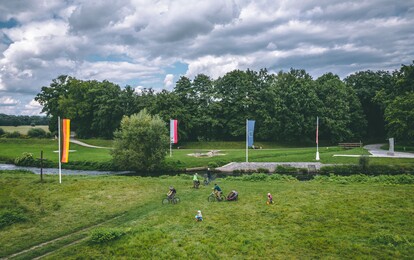 Große Wiesenlandschaft mit Menschen die diese zu Fuß oder mit dem Fahrrad überqueren.