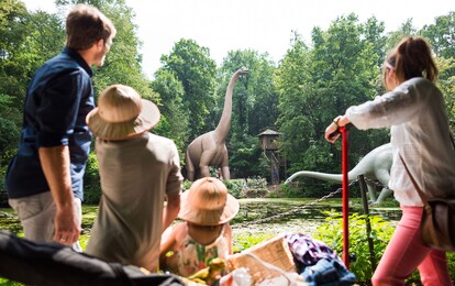 Kinder sehen sich im Saurierpark Kleinwelka Dinosaurierfiguren an. Sie stehen zwischen Bäumen an einem Teich.