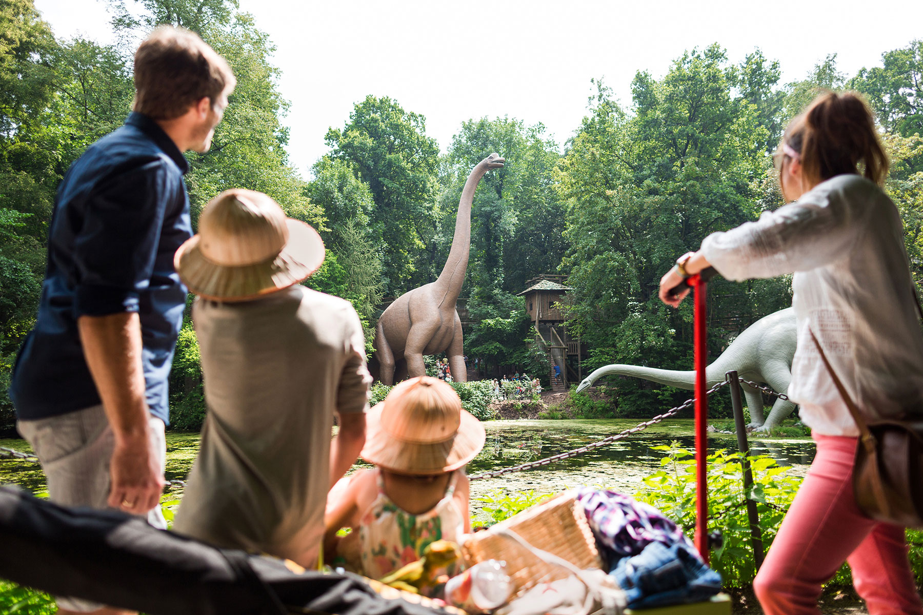 Kinder sehen sich im Saurierpark Kleinwelka Dinosaurierfiguren an. Sie stehen zwischen Bäumen an einem Teich.